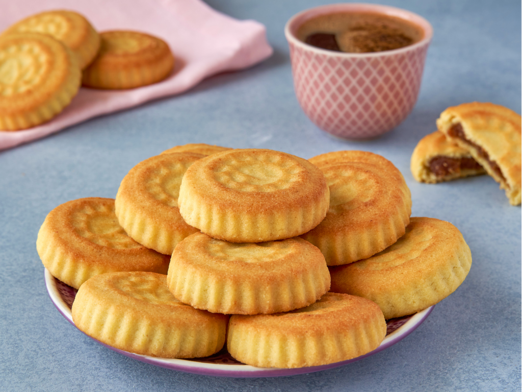 A plate of golden maamoul cookies filled with dates, accompanied by a cup of coffee and a pink napkin in the background.