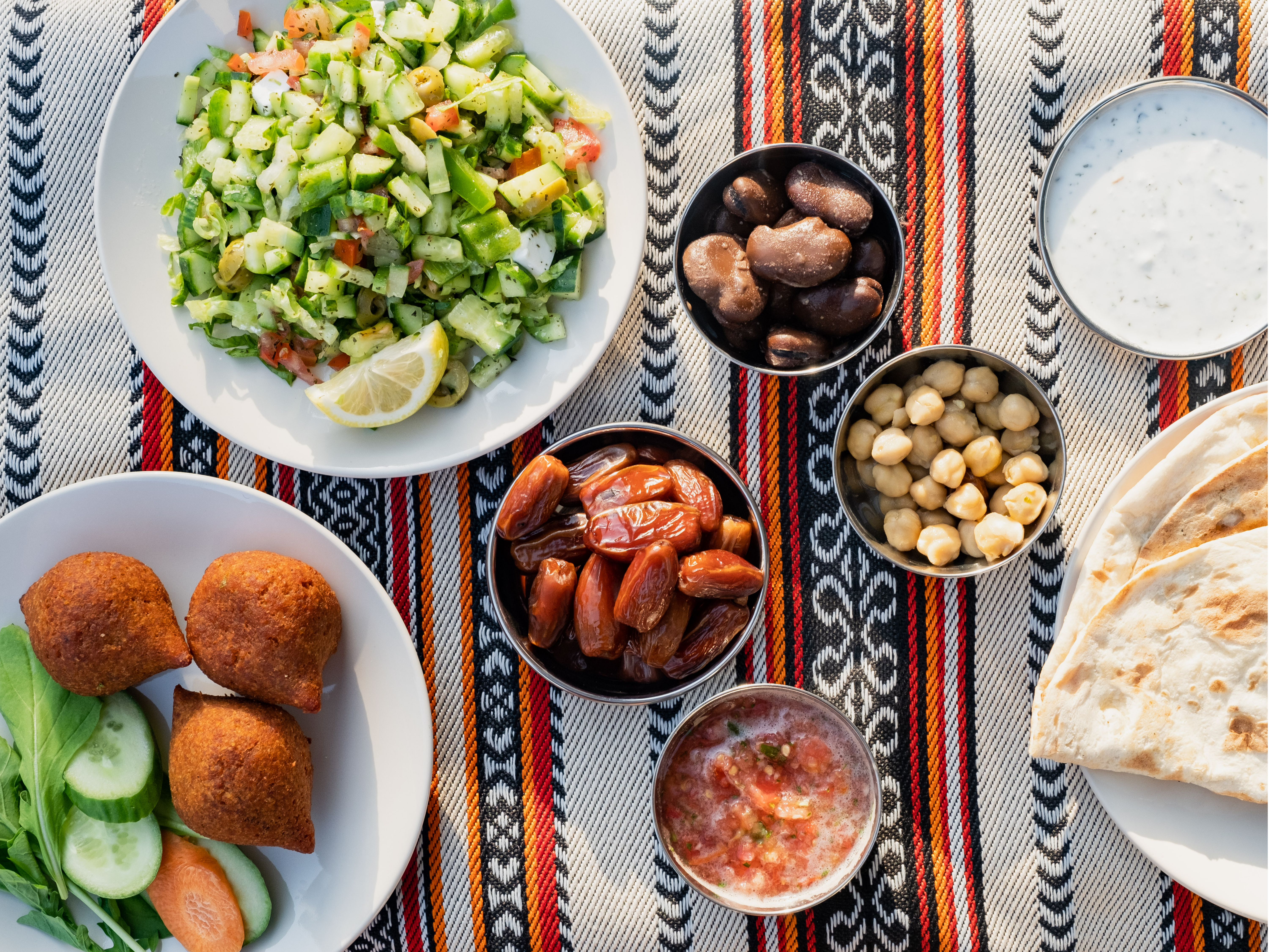 An assortment of Middle Eastern breakfast dishes, including fresh salad, kibbeh, dates, chickpeas, flatbread, and dipping sauces on a patterned textile.