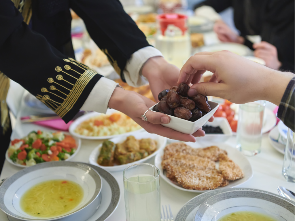 A person in traditional attire offering a bowl of dates to a guest during a festive meal, with a table full of vibrant dishes in the background.