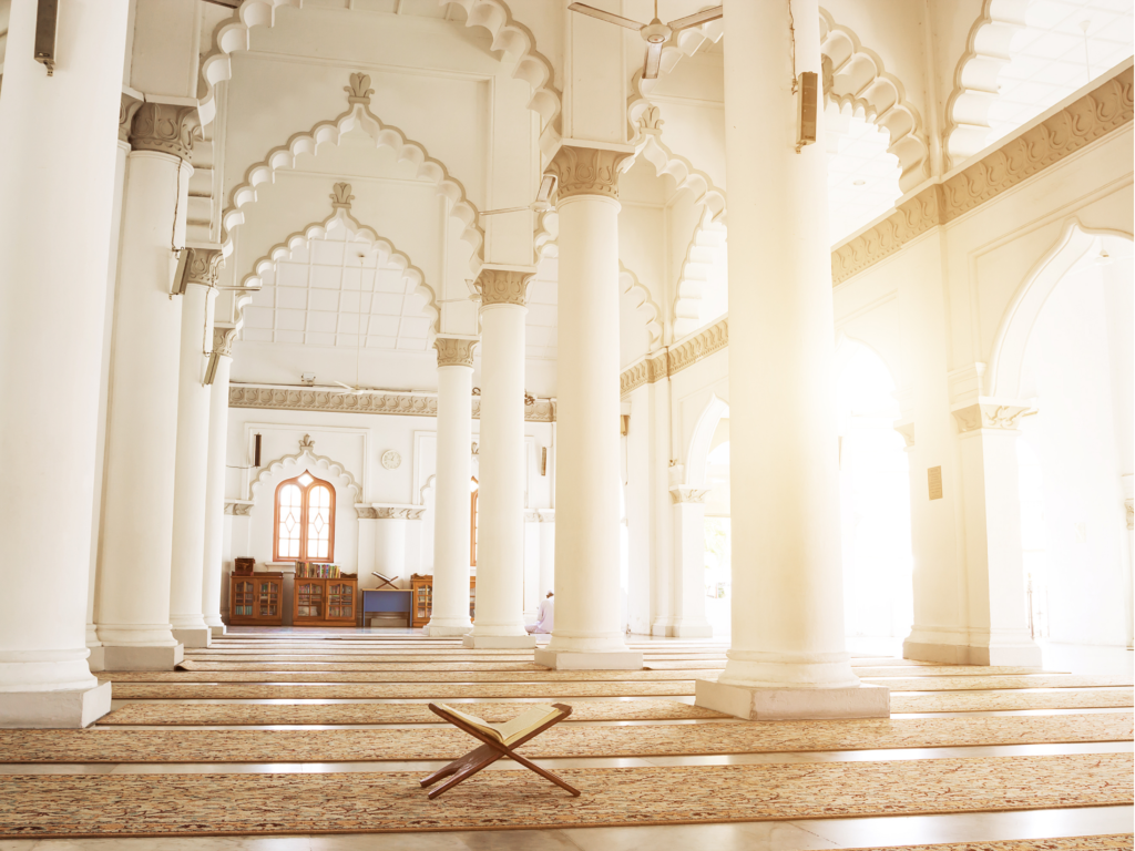 A peaceful mosque interior with tall white columns, arched windows, and a wooden Quran stand bathed in warm sunlight.