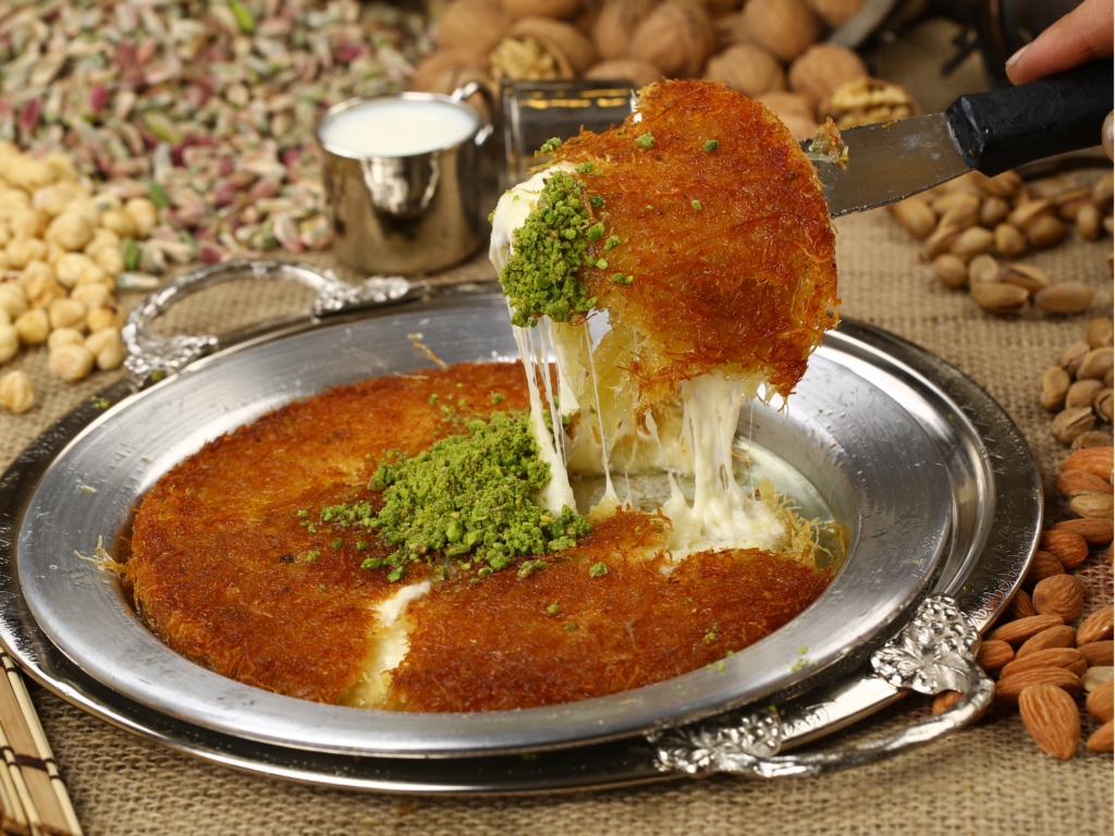 Close-up of a slice of kunafa being lifted from a tray, revealing melted cheese, topped with crushed pistachios, surrounded by nuts and milk.