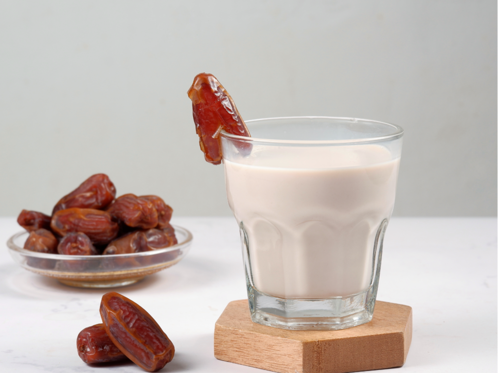 A glass of milk garnished with a fresh date, accompanied by a bowl of dates and a wooden coaster on a white surface.