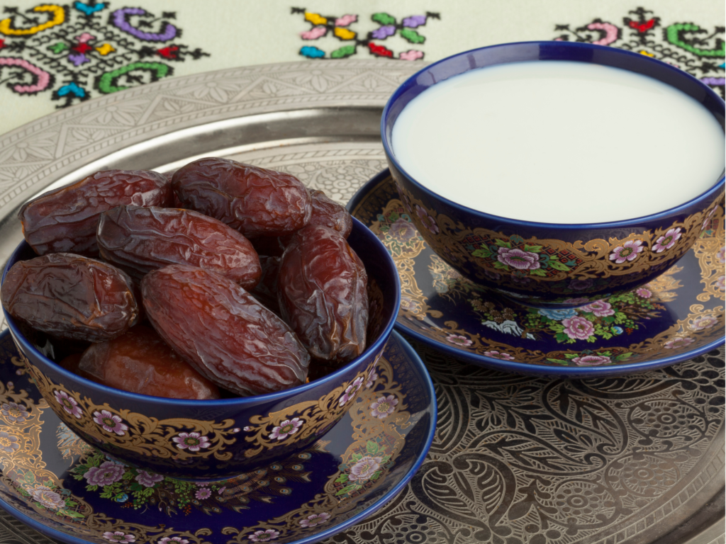 A decorative tray with two beautifully ornate bowls, one filled with fresh dates and the other with milk, set against an embroidered background.