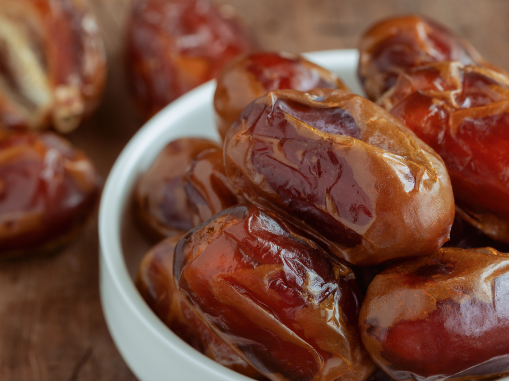 A white bowl filled with glossy, fresh dates, with additional dates blurred in the background on a wooden surface.