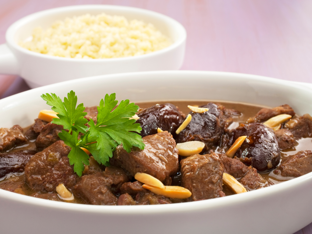 A white dish of braised lamb cooked with dates, toasted almonds, and garnished with parsley, served with a side of couscous.