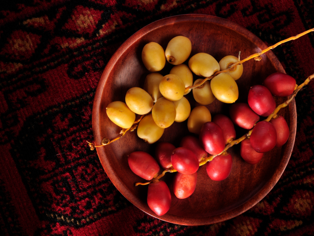 A wooden plate filled with clusters of fresh yellow and red dates, set against a traditional red-patterned fabric.