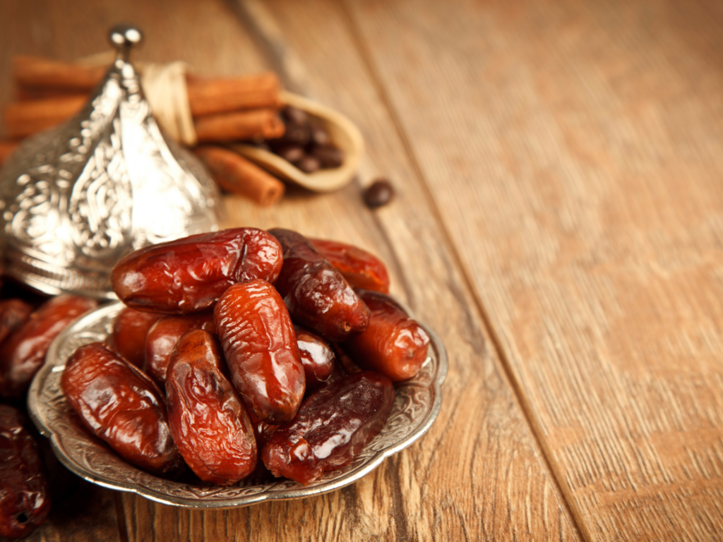 Close-up of glossy dates arranged in an ornate silver dish, accompanied by cinnamon sticks and coffee beans on a wooden surface.