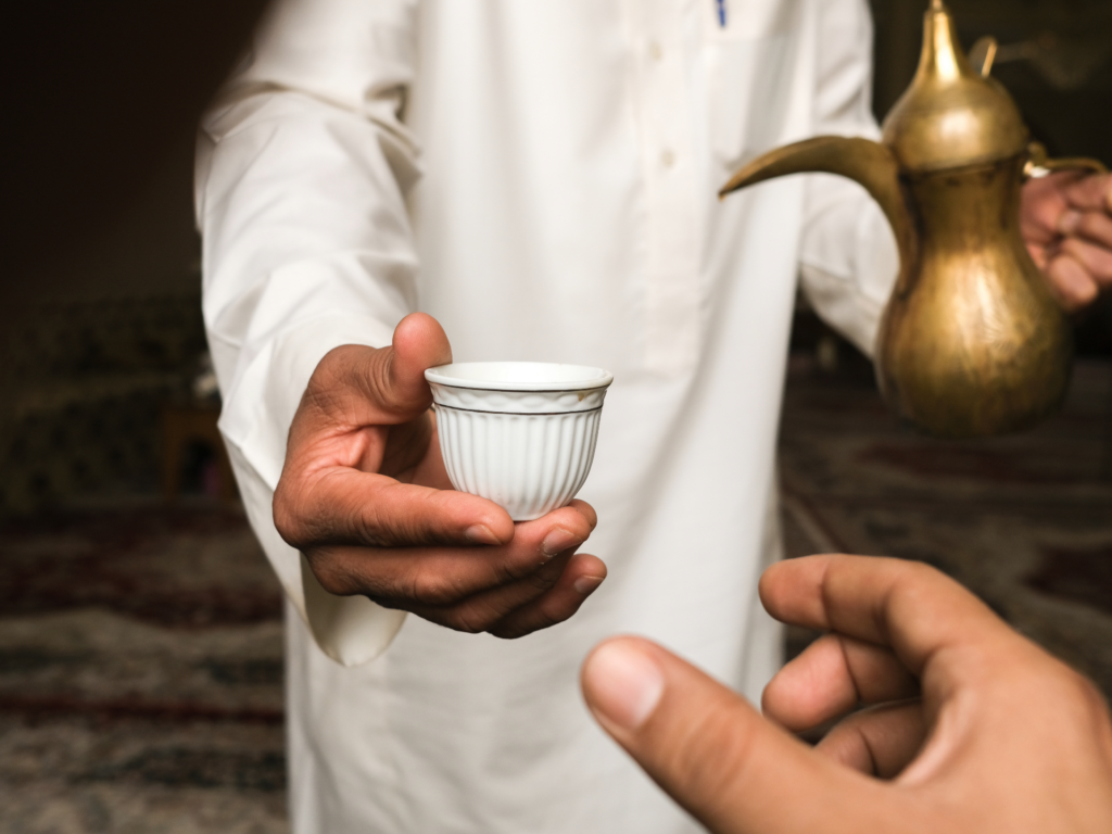 Close-up of a hand offering a small Arabic coffee cup with a golden dallah in the background, symbolizing traditional hospitality.