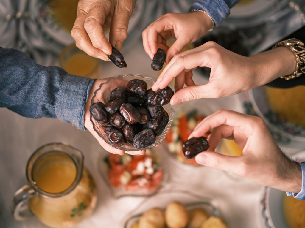 Hands reaching for dates from a bowl during a shared meal, surrounded by traditional food and drinks.